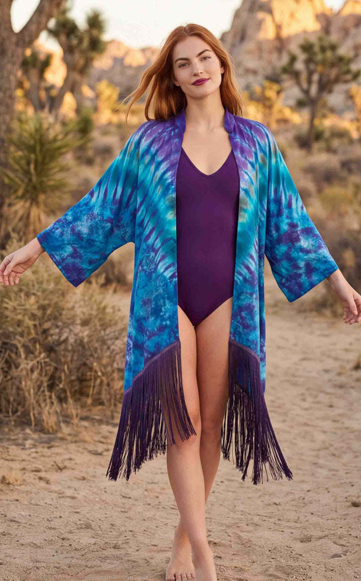 A woman stands barefoot on a sandy desert path wearing a long, open-front fringed cover-up with a blue and purple tie-dye pattern over a purple one-piece swimsuit. Trees and rocky hills are blurred in the background.