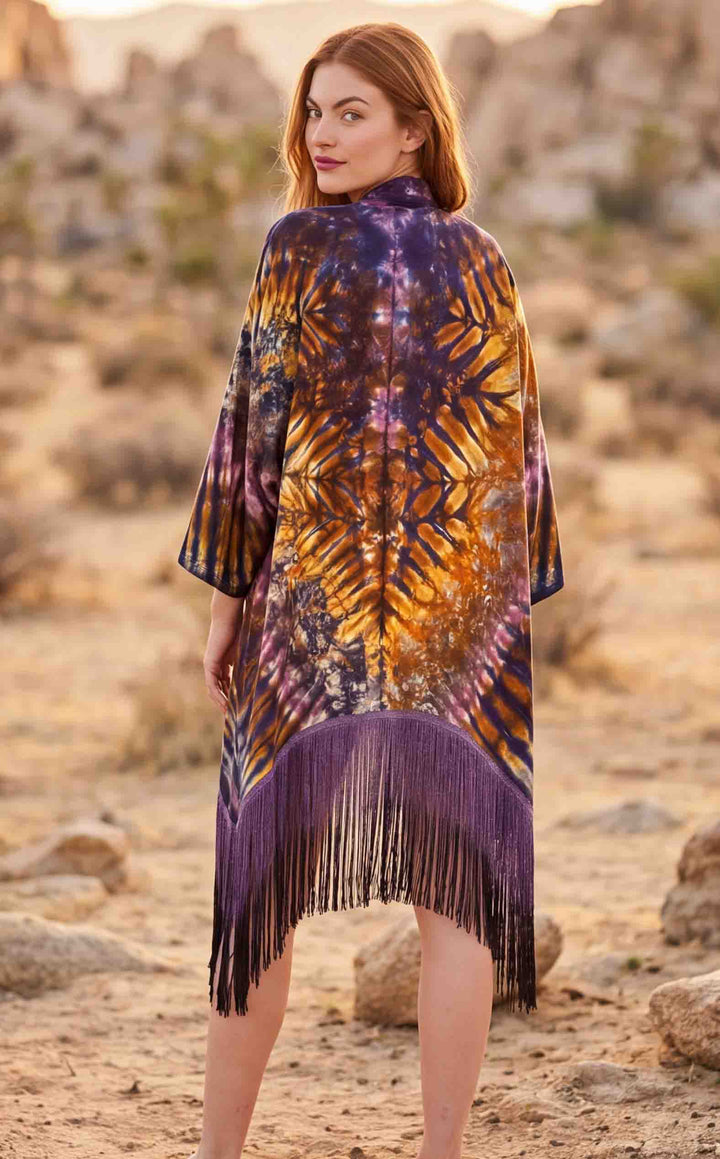 A woman stands in a rocky desert landscape wearing a long, loose fringed poncho or cape with a purple, orange, and yellow tie-dye pattern.