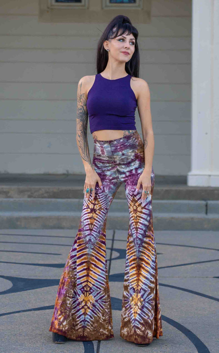 A woman stands outdoors wearing a sleeveless purple crop top and high-waisted, wide-leg flare pants with a multicolor tie-dye pattern in purple, orange, brown, and white.