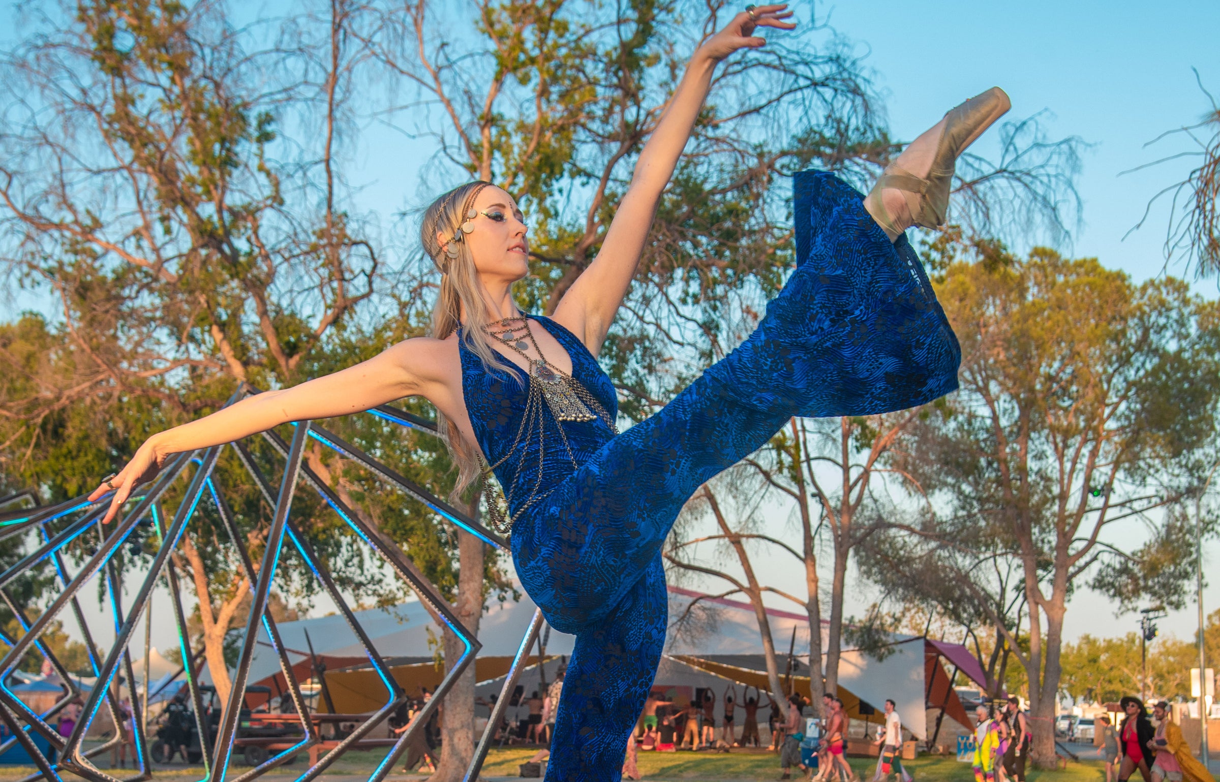 Photo links to New Arrivals. Photo is of a Ballerina wearing a navy velvet jumpsuit adorned in chain headpiece and body chain on torso.  She is doing a high kick. Photo from a music festival