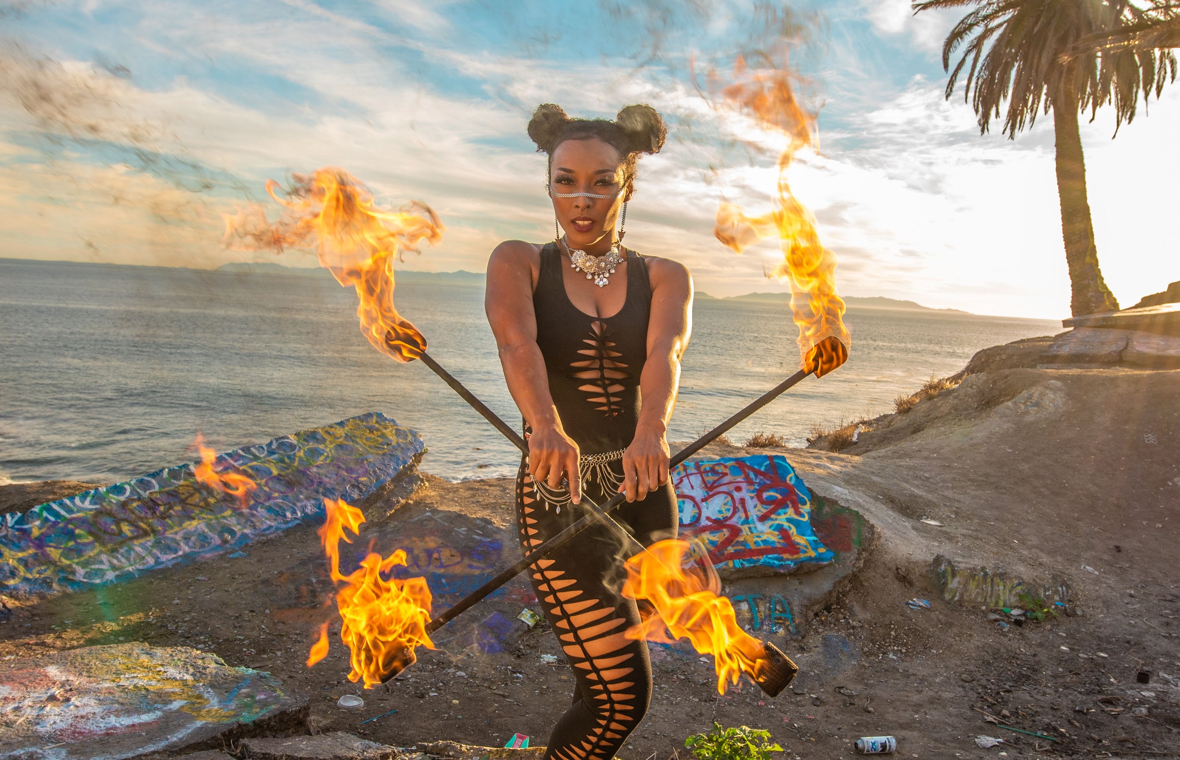 Photo links to Snip Tease Collection which is a Collection of snipped and woven clothing. The image is of a single woman of color who is holding two fire sticks criss-crossed in front of her body. She is wearing a black bamboo cat suit with snip/weaves down front and sides.  She has silver jewelry on her face and neck and belt.  She is standing at sunset on the California Coast