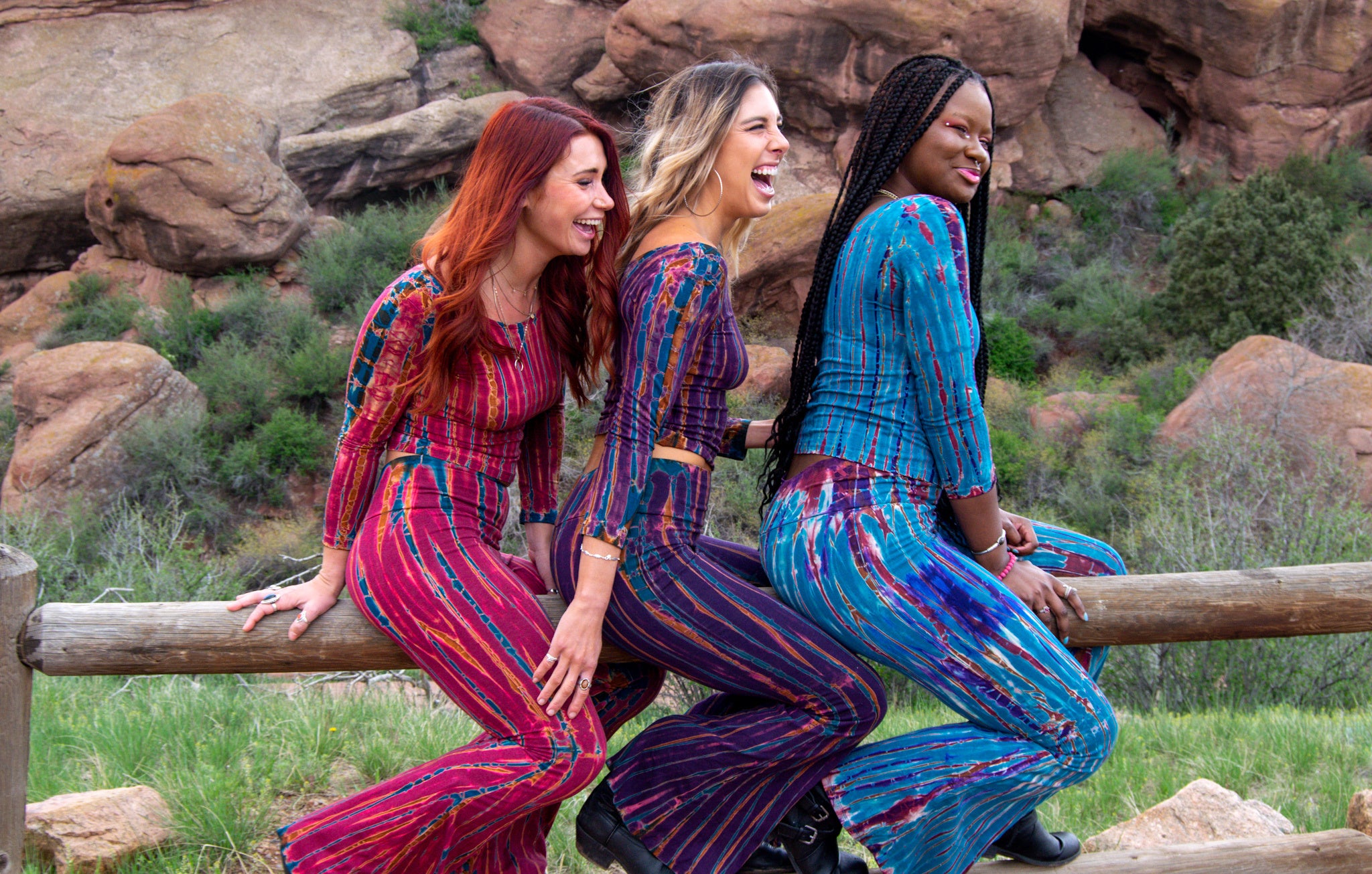 Photo links to Tie Dye Collection. Photo is of three women sitting one in front of the other facing the same direction. They are wearing the same style tie dye in three different colorways. One is maroon with blue/orange detail, next is purple with tan, teal detail and last is teal with purple, tan detail. They are smiling and laughing.  Photo shot at Red Rocks Amphitheater in Colorado.  