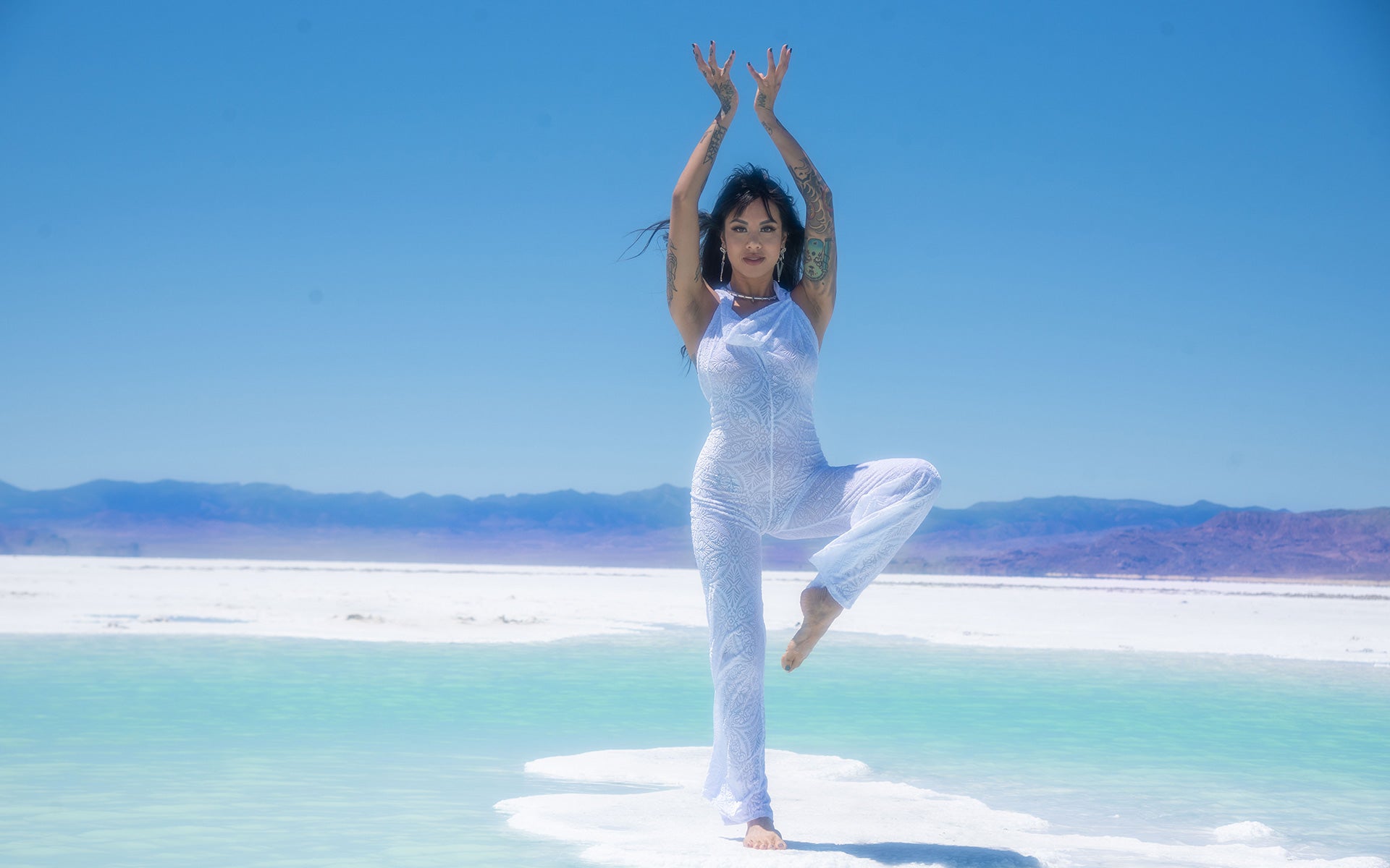 Woman with dark hair wearing a white burnout velvet jumpsuit doing a yoga tree pose with arms above her head. Hair is blowing in the wind.  She is standing in the Great Salt Lake of Utah. It looks like a white sand beach.