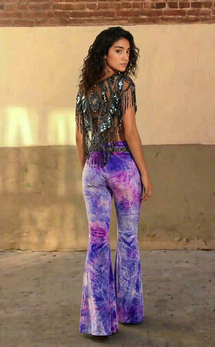 Woman wearing a sequin top and tie-dye flared pants against a beige wall.