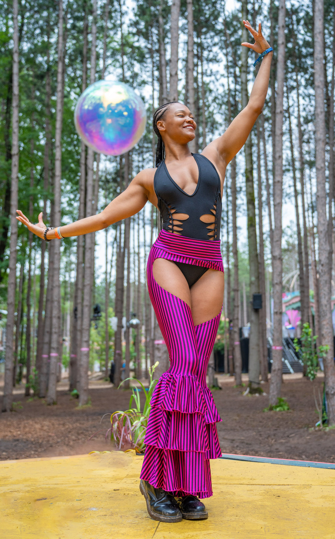 Beautiful model wearing a black snipped leotard under Hot pink & black stripe 3 tiered ruffle  pants. There is a cut out making these chaps. the background is Electric Forest Festival. Forest trees in background & one big bubble orb in the background. 