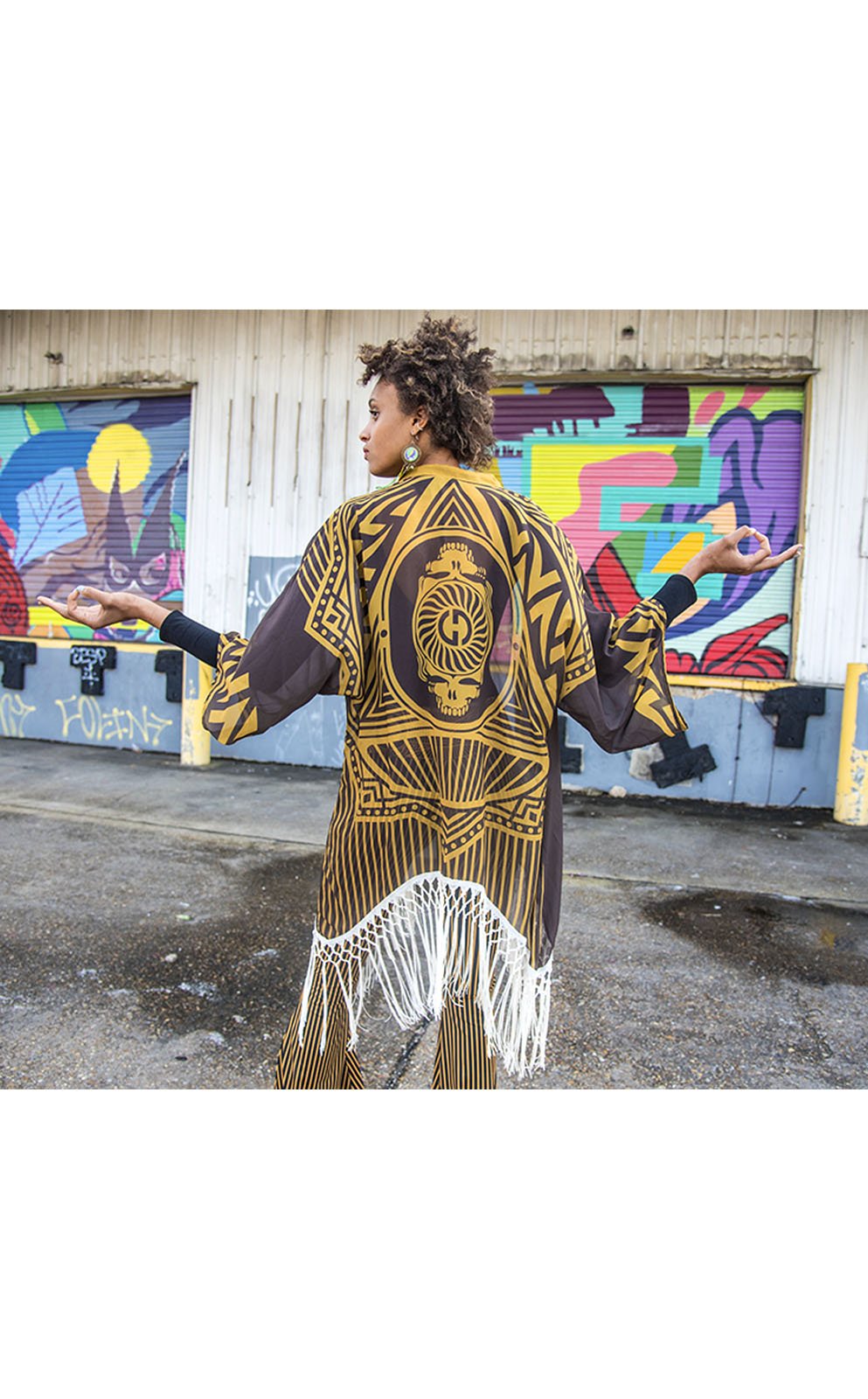 Kimono with long braided cream fringe. The artwork is of a Stealie with stripes based off the 1977 album cover. It is the gold and brown colors mimicking the original album cover. The model is African American and facing backward showing the back of the garment. 