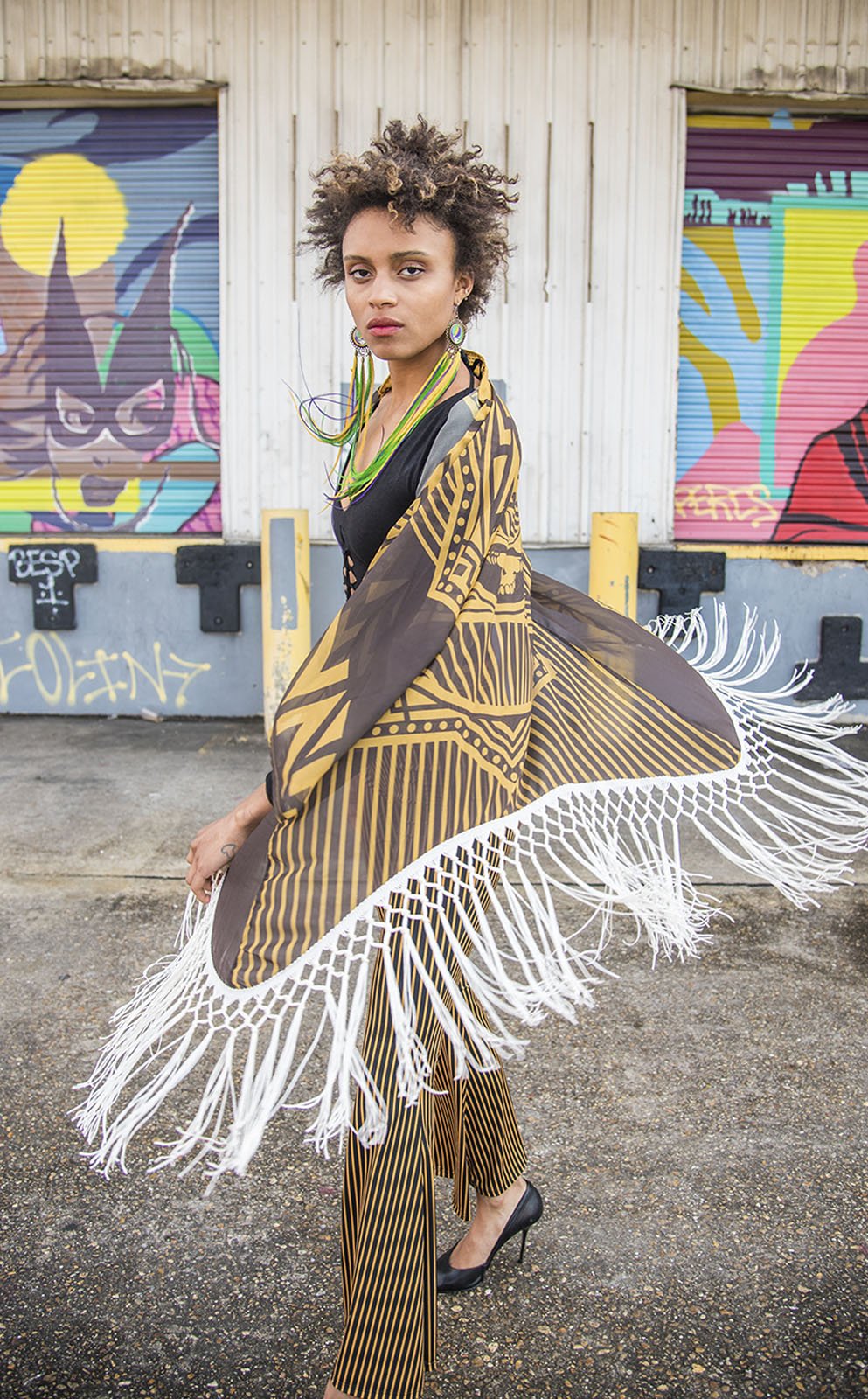 Kimono with long braided cream fringe. The artwork is of a Stealie with stripes based off the 1977 album cover. It is the gold and brown colors mimicking the original album cover. This is a close up shot of the models top halfe with the fringe in motion. Model has serious face. She is African American woman with short hair. 