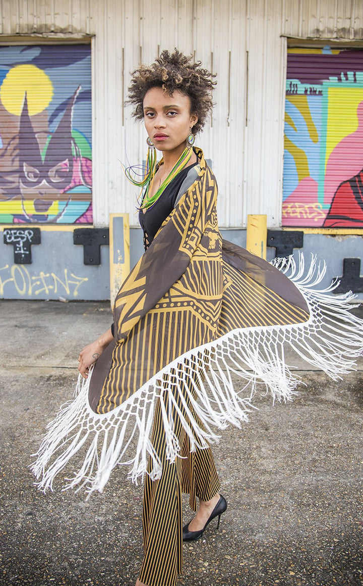 Kimono with long braided cream fringe. The artwork is of a Stealie with stripes based off the 1977 album cover. It is the gold and brown colors mimicking the original album cover. This is a close up shot of the models top halfe with the fringe in motion. Model has serious face. She is African American woman with short hair. 