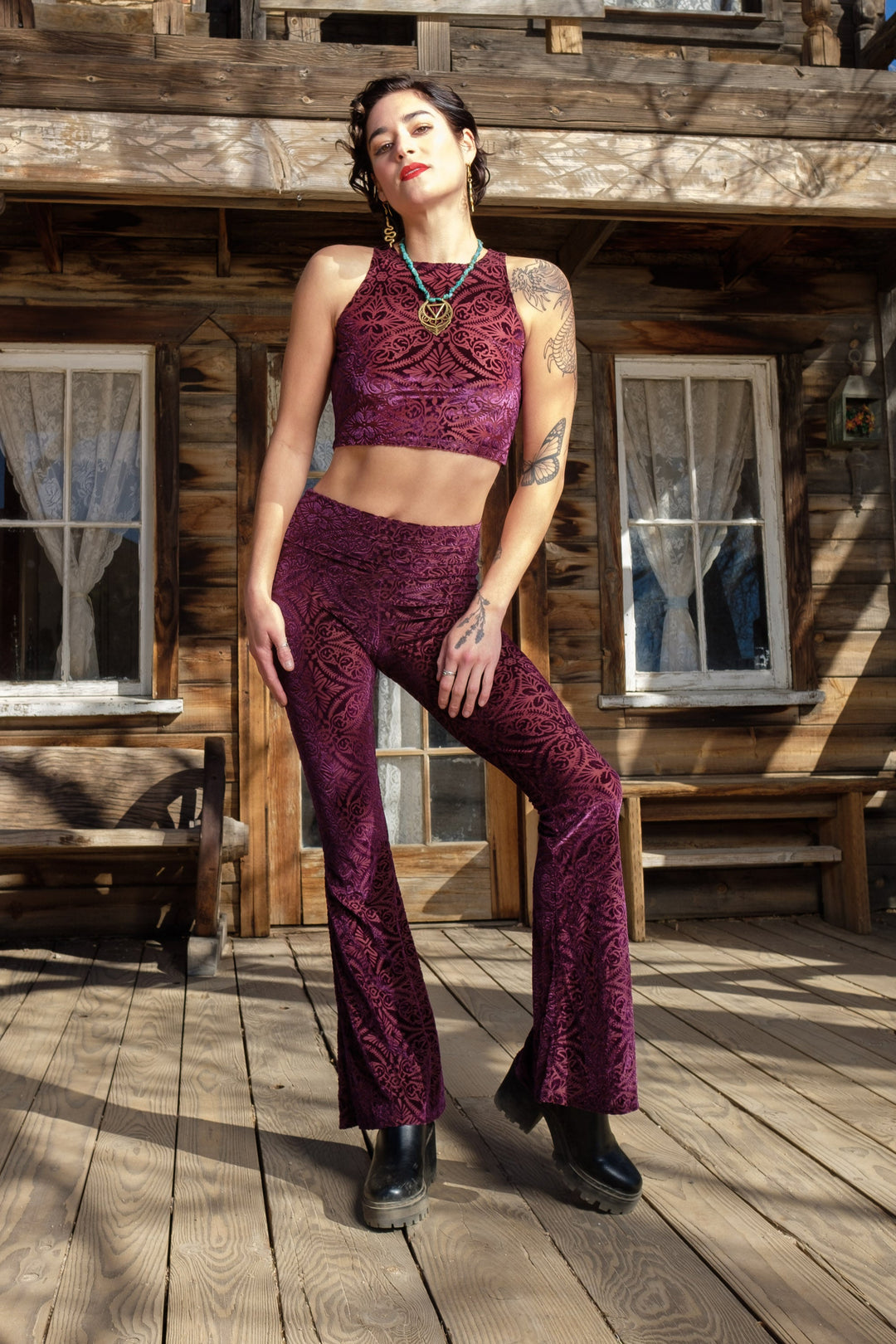 A person wearing maroon burnout velvet slim bell pants with a wider waistband, standing in front of an old wooden building.
