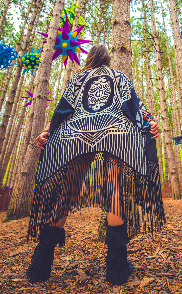 Kimono with long braided black fringe. The artwork is of a Stealie with stripes based off the 1977 album cover. It is the black and white. The model facing backwards showing the back of the garment.  This image is shot from below looking up at the model. 