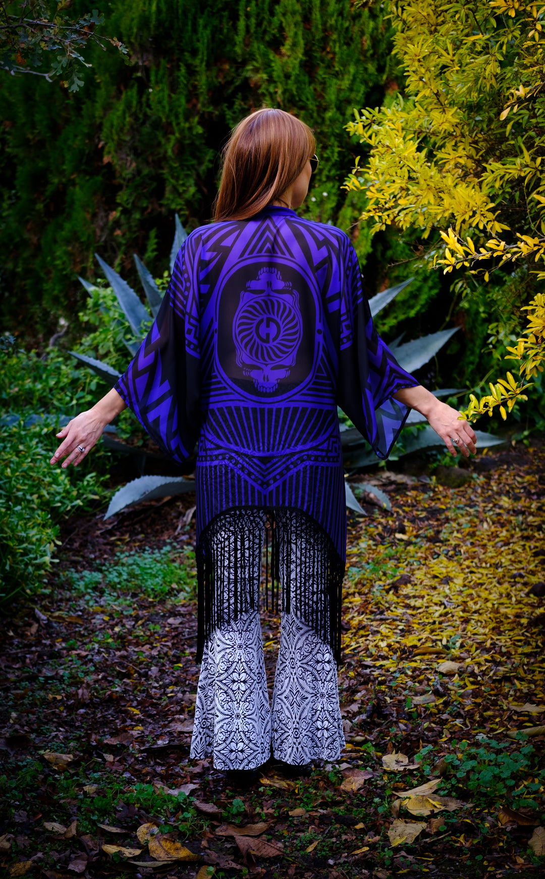 Kimono with long braided black fringe. The artwork is of a Stealie with stripes based off the 1977 album cover.  The print is black and purple. The model is faced backward not showing any of her face. 
 You can see the full backside of the garment. 
