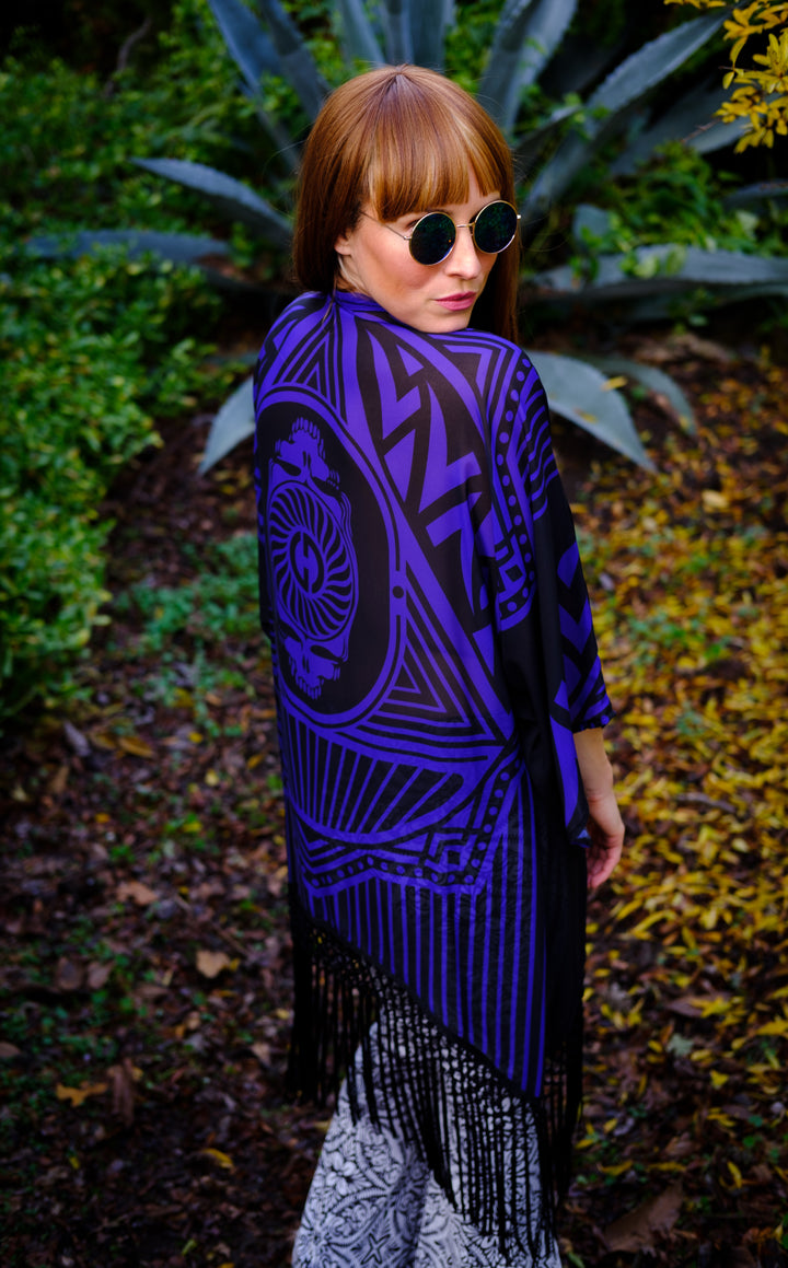 Kimono with long braided black fringe. The artwork is of a Stealie with stripes based off the 1977 album cover.  The print is black and white. The model is wearing sunglasses and faced to the side so you can see most of the back of the garment. 