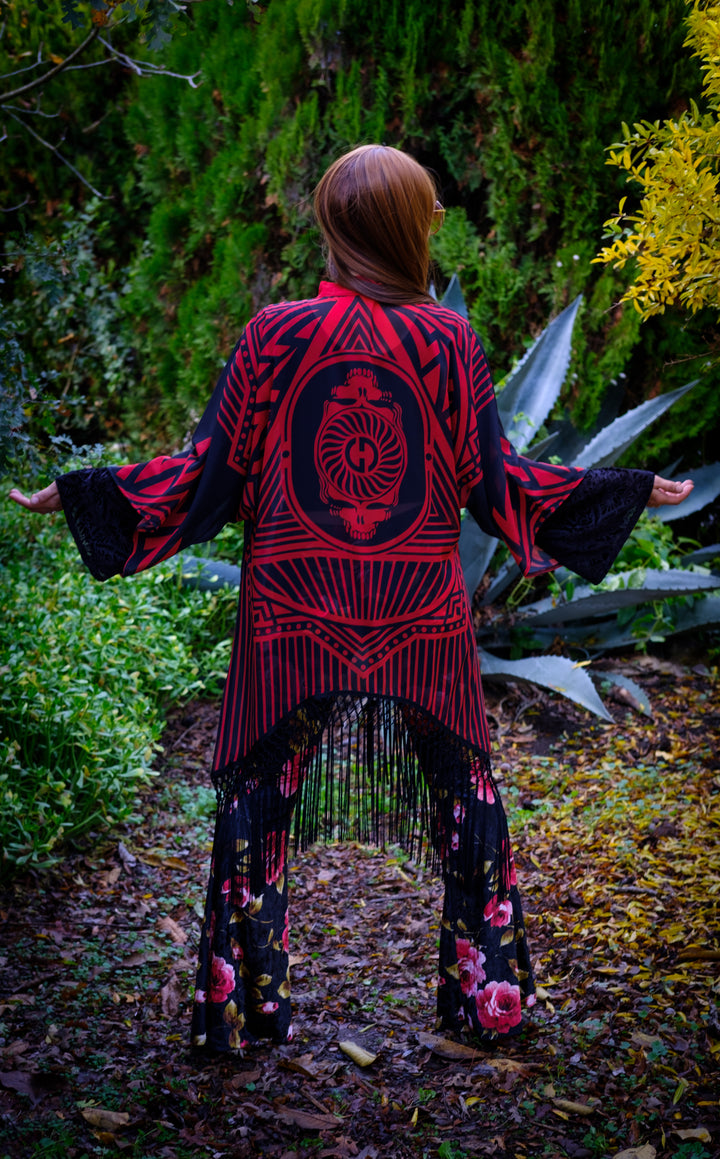 Kimono with long braided black fringe. The artwork is of a Stealie with stripes based off the 1977 album cover.  The print is black and red. The model is faced backward not showing any of her face. 
 You can see the full backside of the garment. 